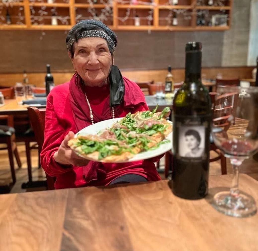 Luisa holding a plate in the dining room, beside a bottle of the house-label wine bearing her portrait